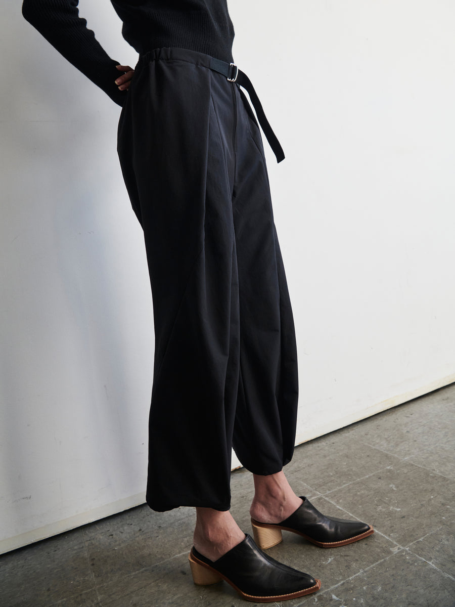 Wearing the Wrap Akeo Pant by Zero + Maria Cornejo, a person is seen standing in a minimalist room with a gray floor and white walls. The outfit comprises black trousers featuring an adjustable belt and black heeled shoes, paired with a long-sleeve dark top made from technical-stretch woven fabric. Their hands rest comfortably in the trousers' pockets.