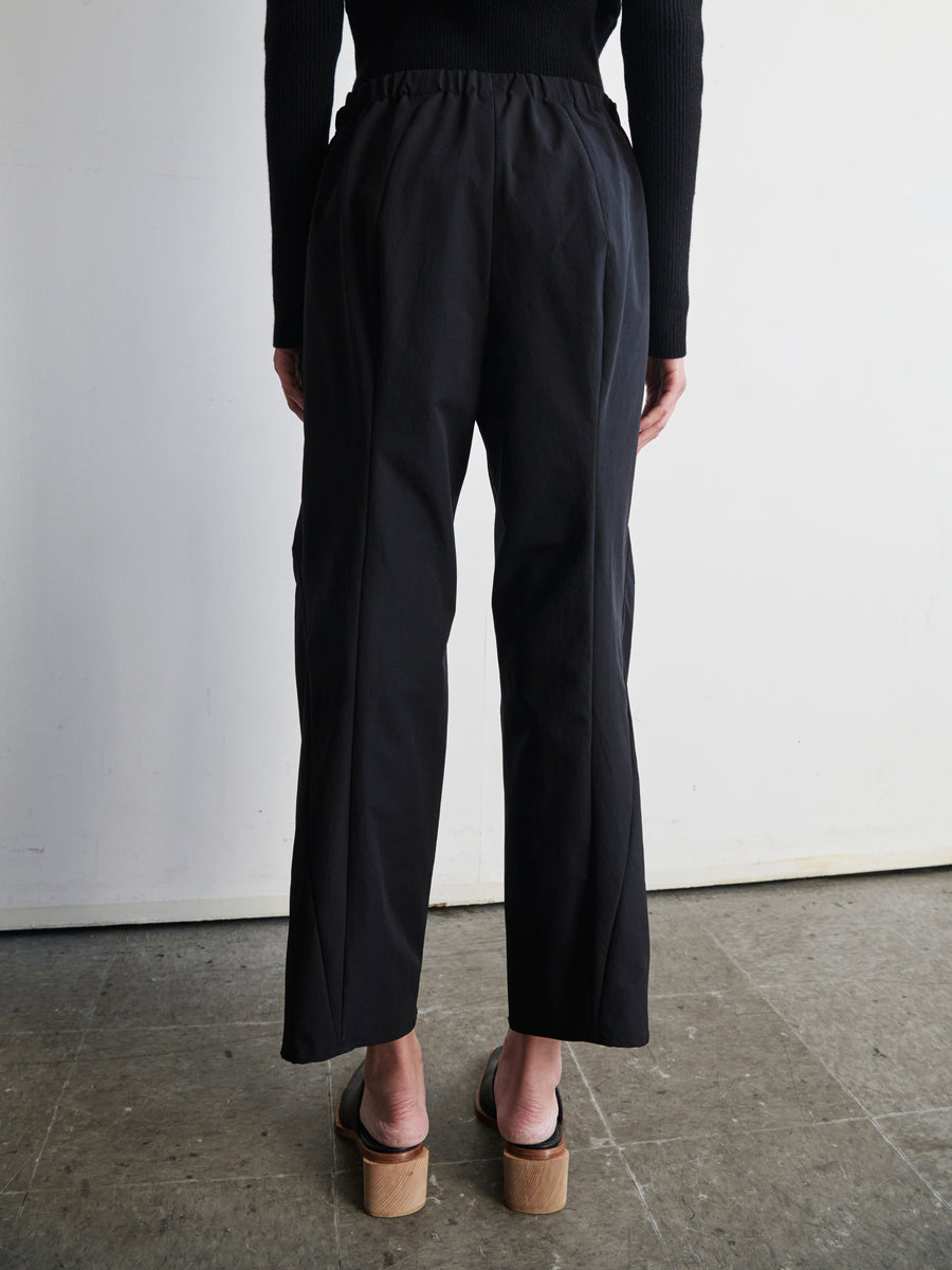 A person stands with their back to the camera, highlighting a black long-sleeve top and the Wrap Akeo Pant by Zero + Maria Cornejo, crafted from eco-friendly wrinkle-resistant fabric, complemented by wooden platform sandals. The minimalist setting features a plain white wall and gray floor.