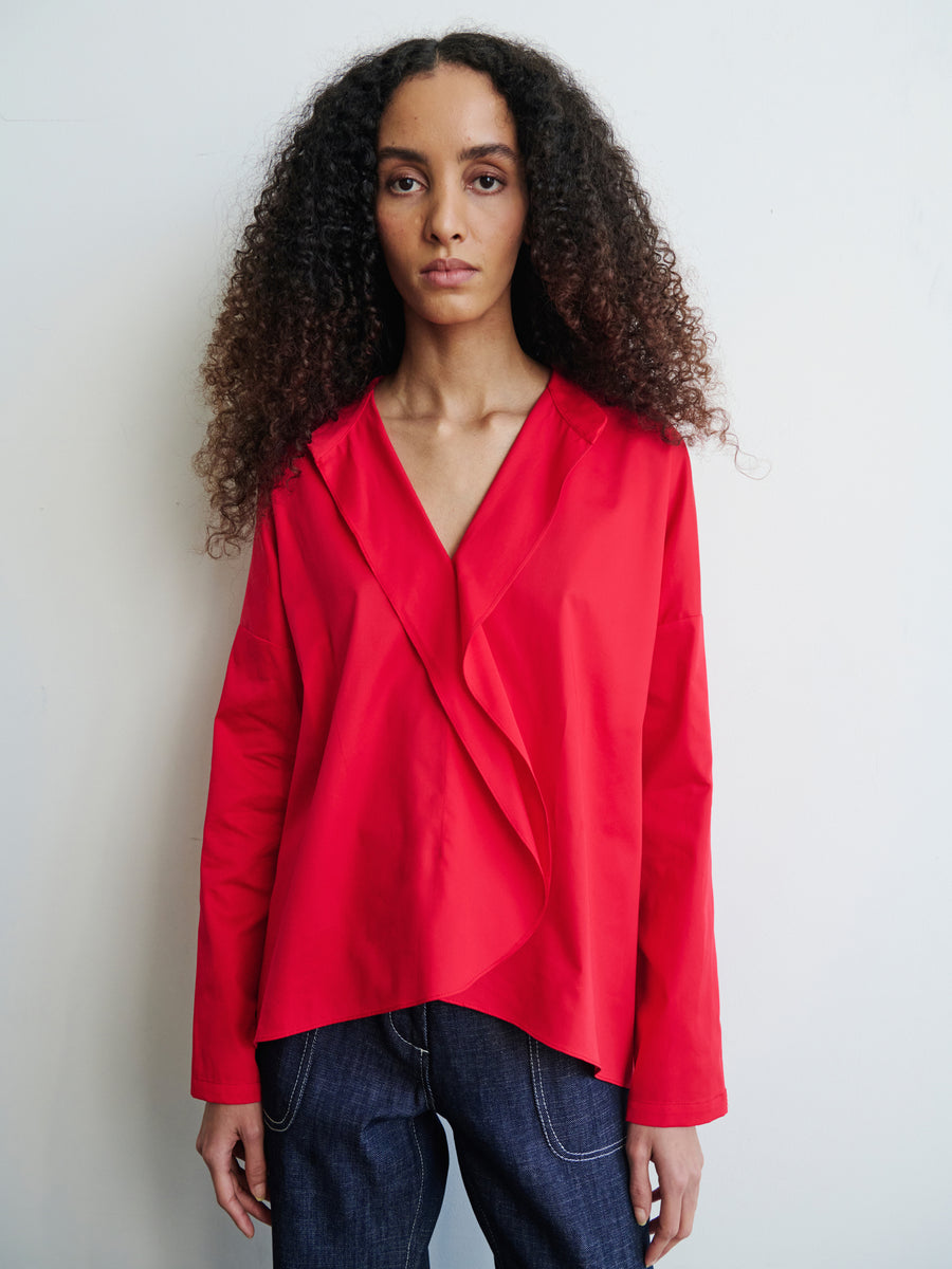 A person with long curly hair wears the Long-Sleeved Fin Gaban Shirt by Zero + Maria Cornejo, paired with dark jeans, against a plain white background. They have a neutral expression and are facing forward.