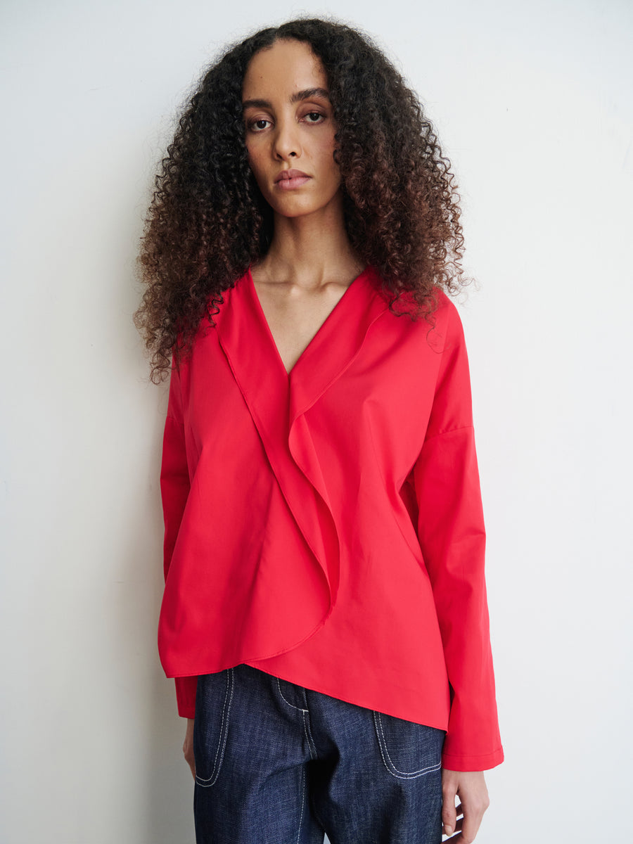 Against a plain white background, an individual with long curly hair is seen wearing the Zero + Maria Cornejo Long-Sleeved Fin Gaban Shirt, which features a bright red color, stand collar, and distinctive ruffle detail. They pair it with dark blue pants and maintain a calm expression.