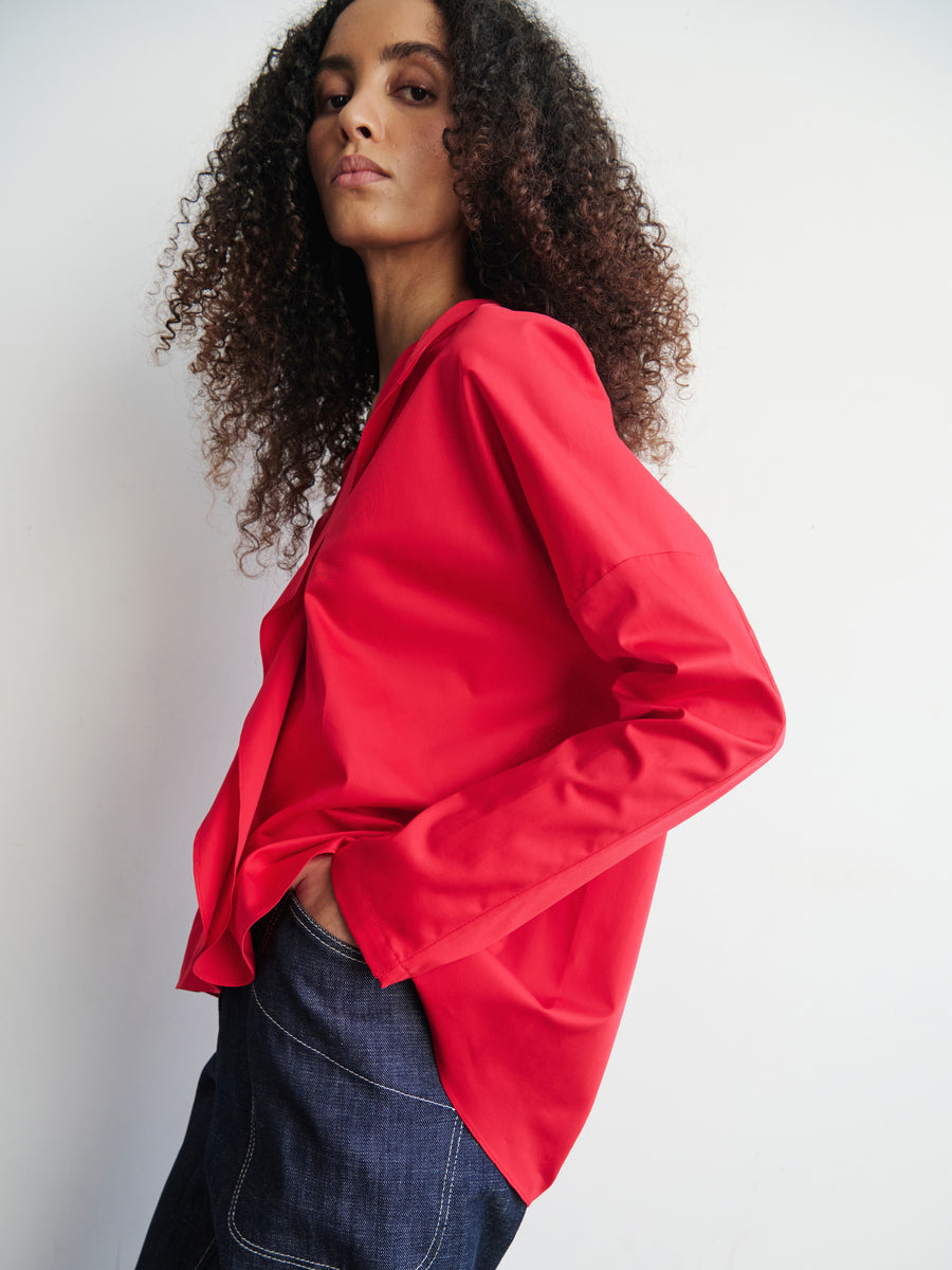 A person with curly hair, confidently poses against a plain white background wearing a bright red Long-Sleeved Fin Gaban Shirt by Zero + Maria Cornejo and dark denim pants, one hand in their pocket as they look directly at the camera.