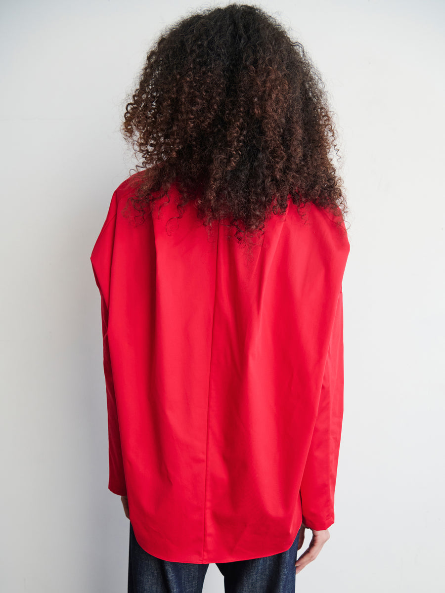 Rear view of a person with curly hair wearing a bright red Long-Sleeved Fin Gaban Shirt by Zero + Maria Cornejo and dark jeans, standing against a light-colored background.