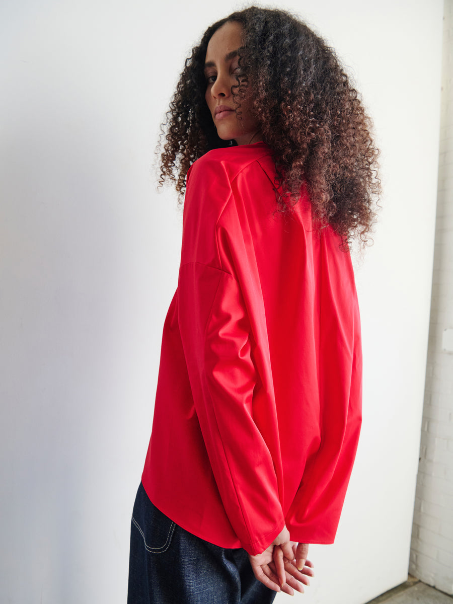 A person with curly hair wearing a vibrant red Long-Sleeved Fin Gaban Shirt by Zero + Maria Cornejo and dark pants stands against a white wall, looking over their shoulder.