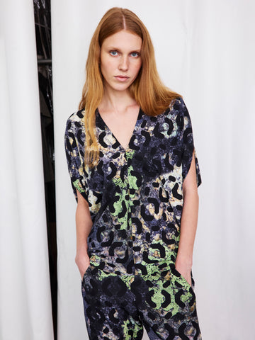 A woman with long, straight, light brown hair stands before white curtains wearing the Zero + Maria Cornejo Kio Top and matching pants in a dark blue, green, and beige abstract archival print, with her hands in her pockets.
