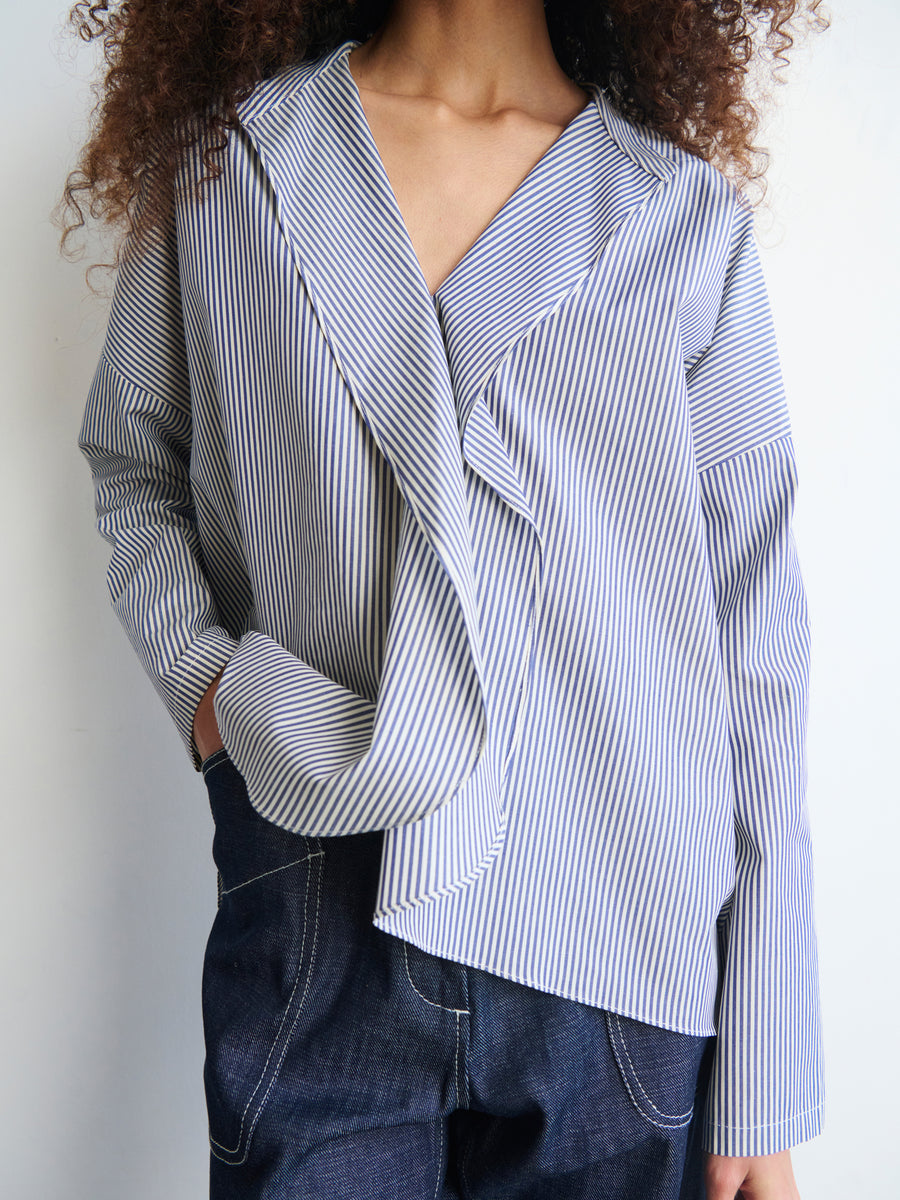 A person wearing a stylish Long-Sleeved Fin Gaban Shirt from Zero + Maria Cornejo, featuring unique draping across the front for an oversized fit, paired with dark denim jeans. They stand against a plain background with one hand in their pocket, and their curly hair is visible.