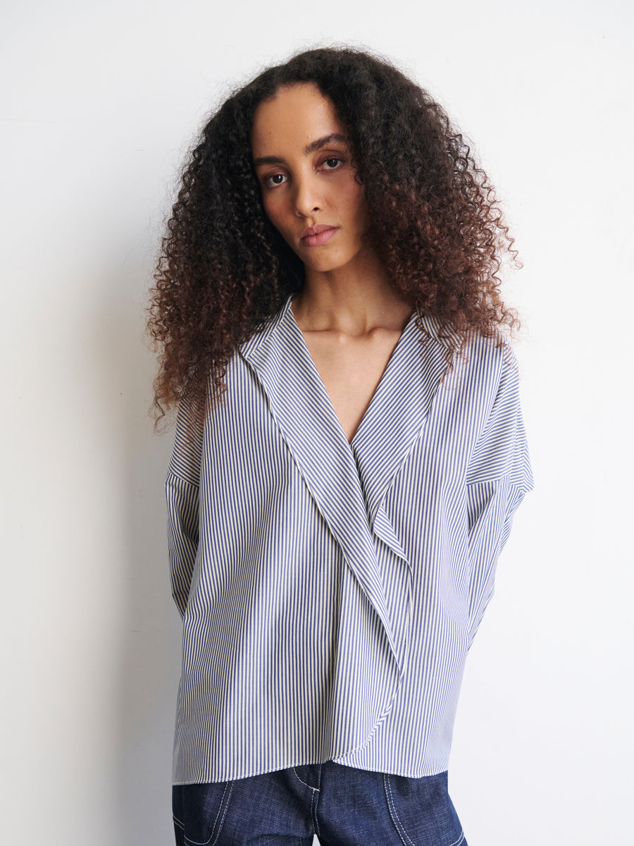 A person with curly hair poses against a plain white backdrop, wearing an oversized Long-Sleeved Fin Gaban Shirt by Zero + Maria Cornejo along with dark pants. They look thoughtfully at the camera with a neutral expression.