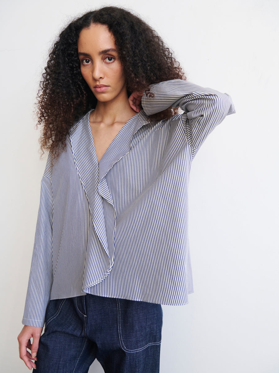 A person with long curly hair stands against a plain background, wearing an oversized Long-Sleeved Fin Gaban Shirt by Zero + Maria Cornejo and dark jeans. They have a contemplative expression, with one hand raised to touch their hair.