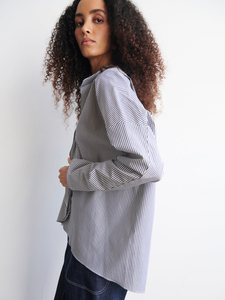 A person with long, curly hair poses against a white background, wearing an oversized Long-Sleeved Fin Gaban Shirt by Zero + Maria Cornejo in blue and white stripes, paired with dark jeans. They gaze to the side with a neutral expression.