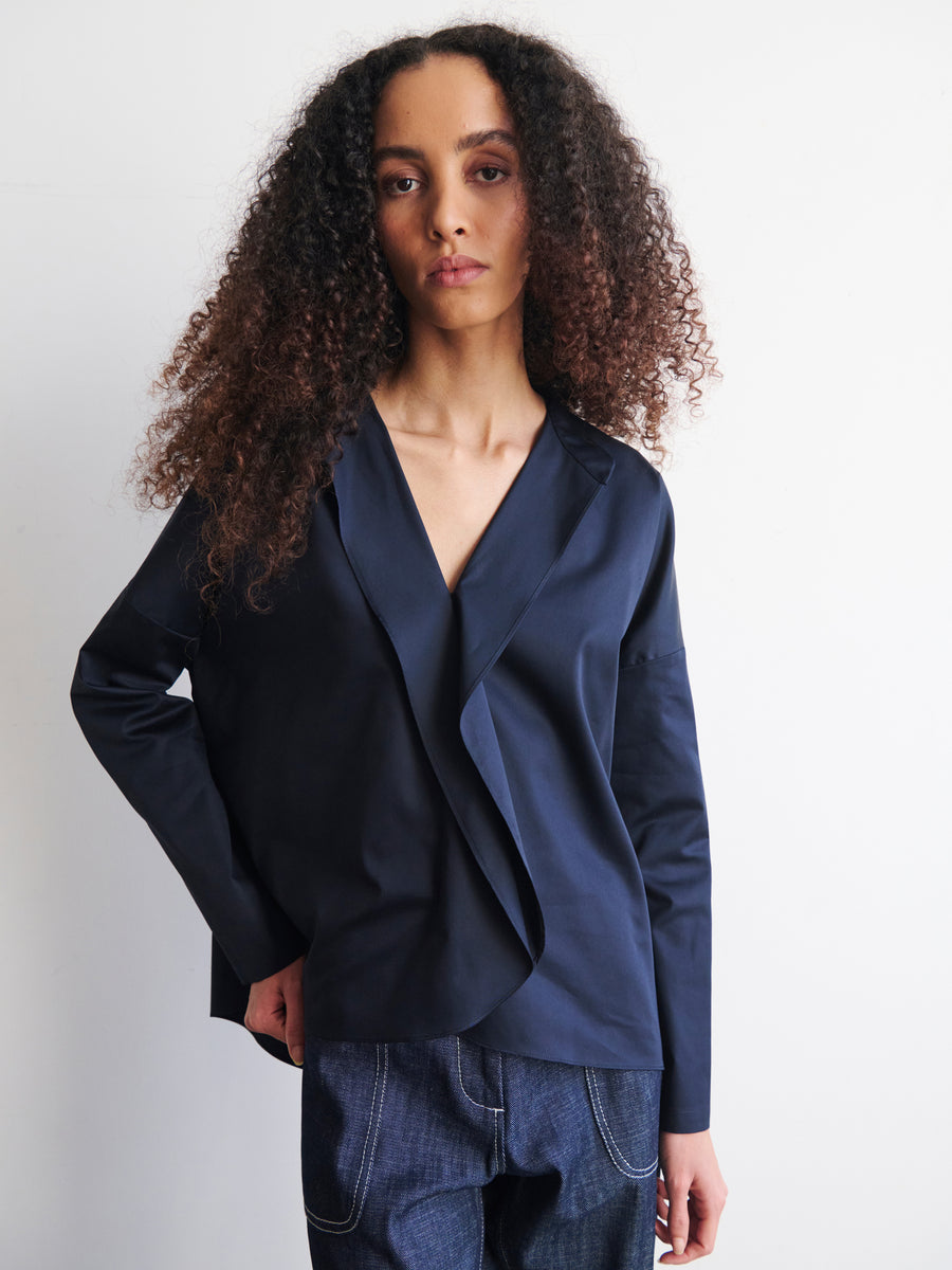 A person with long, curly hair is wearing the Long-Sleeved Fin Gaban Shirt by Zero + Maria Cornejo, which features a stylish draped ruffle and stand collar. They have paired it with dark pants and are standing against a plain white background, looking directly at the camera with a calm expression.