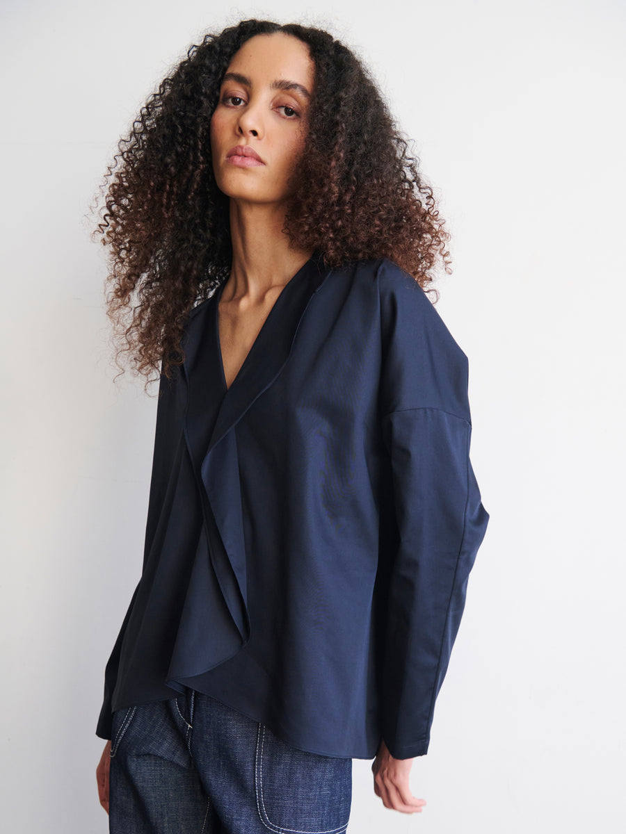 A person with curly hair dons a navy Long-Sleeved Fin Gaban Shirt by Zero + Maria Cornejo, showcasing draped ruffle details and a layered design. They pair it with dark jeans and stand against a plain white background, exuding a relaxed and confident expression.