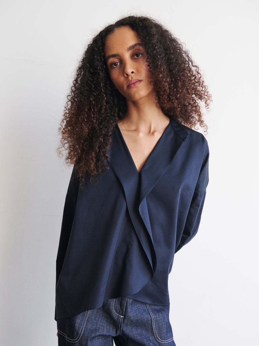 A person with long curly hair is wearing a Long-Sleeved Fin Gaban Shirt by Zero + Maria Cornejo, standing against a plain white background with a relaxed and neutral expression.