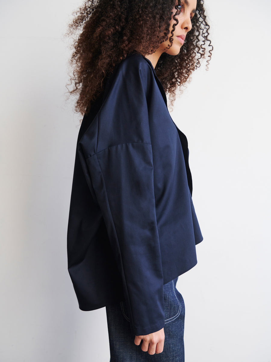 A person with curly hair is wearing a dark blue jacket over Zero + Maria Cornejo's Long-Sleeved Fin Gaban Shirt, which features a stand collar, paired with dark jeans, standing in profile against a plain white background.