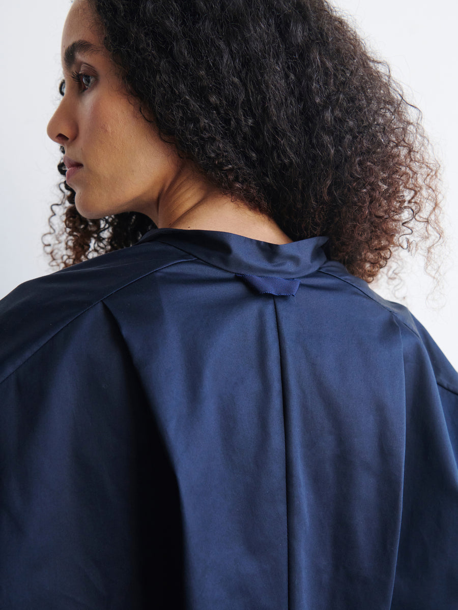 A person with curly hair is turned to the side, wearing a Long-Sleeved Fin Gaban Shirt in dark blue cotton broadcloth by Zero + Maria Cornejo, showcasing its unique back design. The focus is on the texture and structure of the clothing against a plain white background.