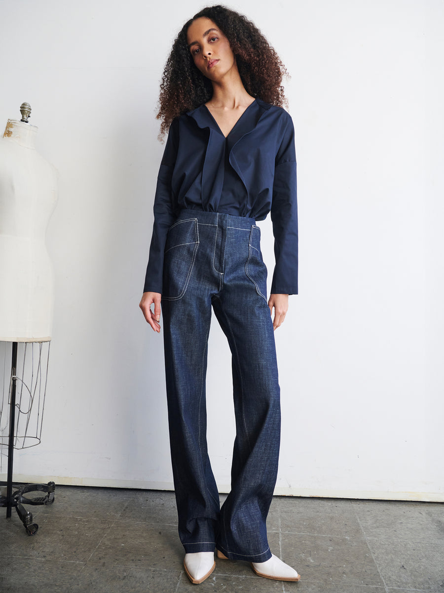 A person with long curly hair is posing in a stylish dark blue top and Eda Pant from Zero + Maria Cornejo. They are standing next to a dress form in a minimalist room with a neutral-colored wall and floor.