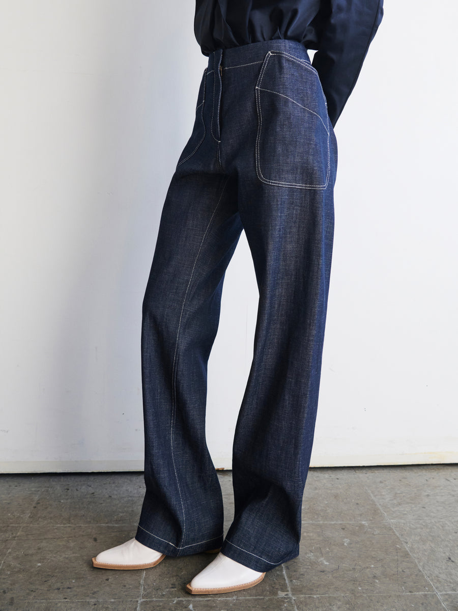 A person wearing the Eda Pant by Zero + Maria Cornejo, featuring wide-leg organic denim with white stitching, and cream-colored pointed shoes stands against a light-colored wall. The ensemble is completed with a dark top, and the floor beneath has a mottled grayish hue.