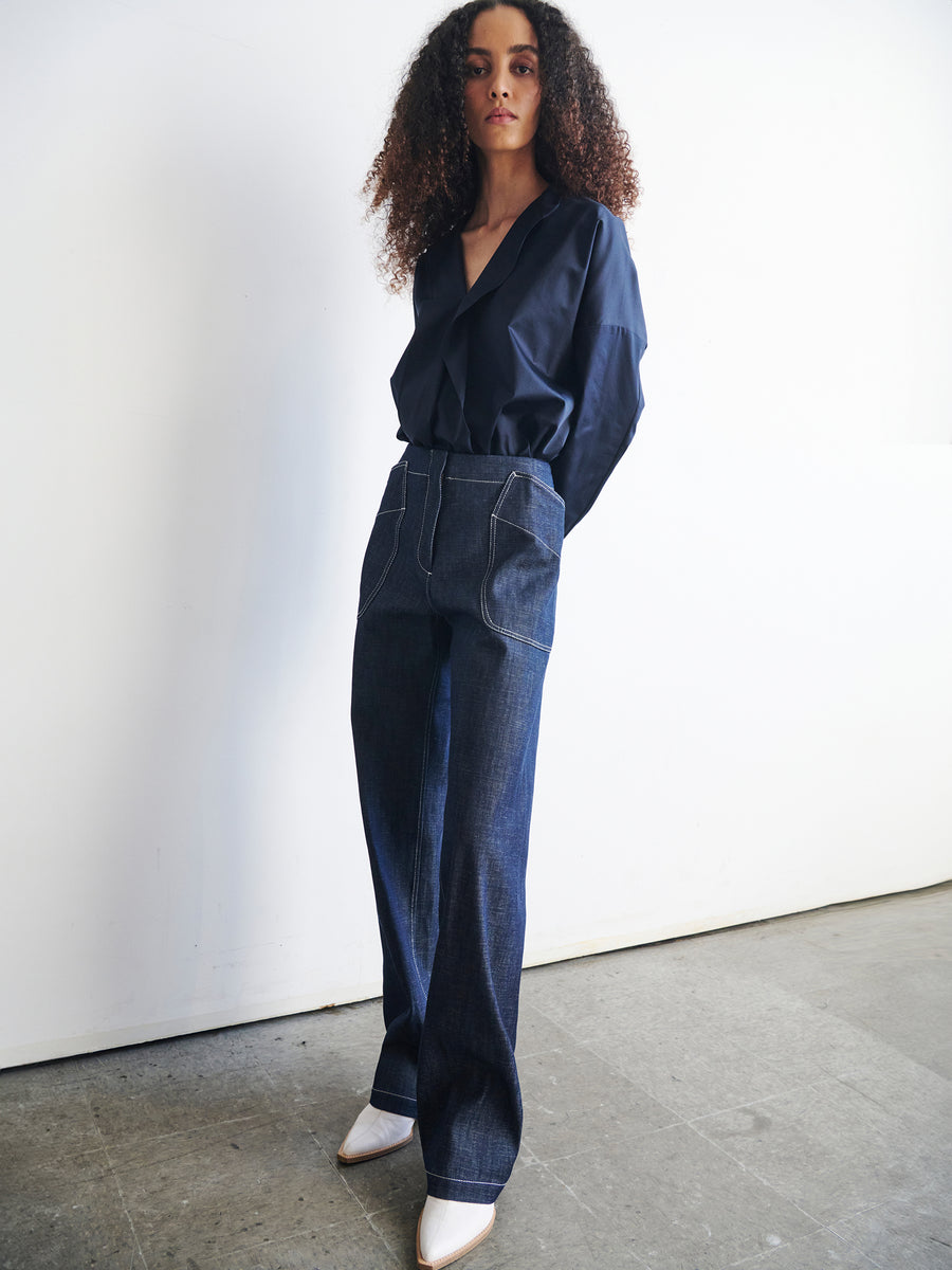 A person with curly hair stands confidently wearing a navy blouse and the high-waisted, wide-leg Eda Pant from Zero + Maria Cornejo. They have white pointed shoes and are posed against a simple, light-colored background.