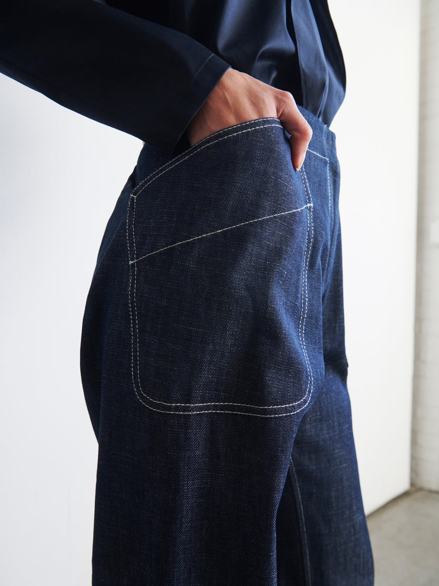 Close-up of a person wearing Zero + Maria Cornejo's Eda Pant, featuring visible white stitching. They are standing in a slightly turned position with their hand resting in the front pocket of these Cradle 2 Cradle Certified jeans. The person also wears a dark-colored shirt against a plain light wall.