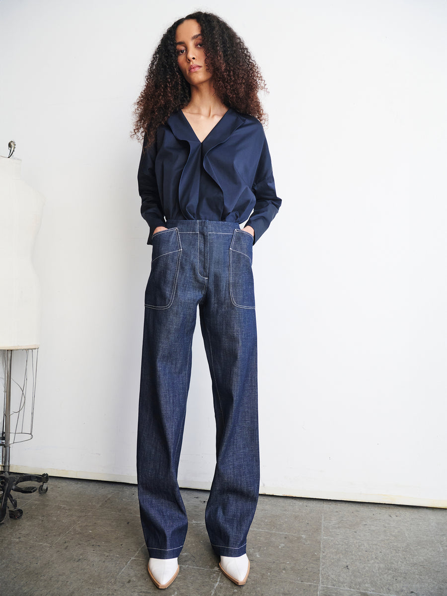 A person with curly hair stands against a plain white wall, wearing a navy blouse and the Eda Pant by Zero + Maria Cornejo with hands in pockets. A dress form is partially visible to the left. They have white pointed shoes and are looking at the camera.