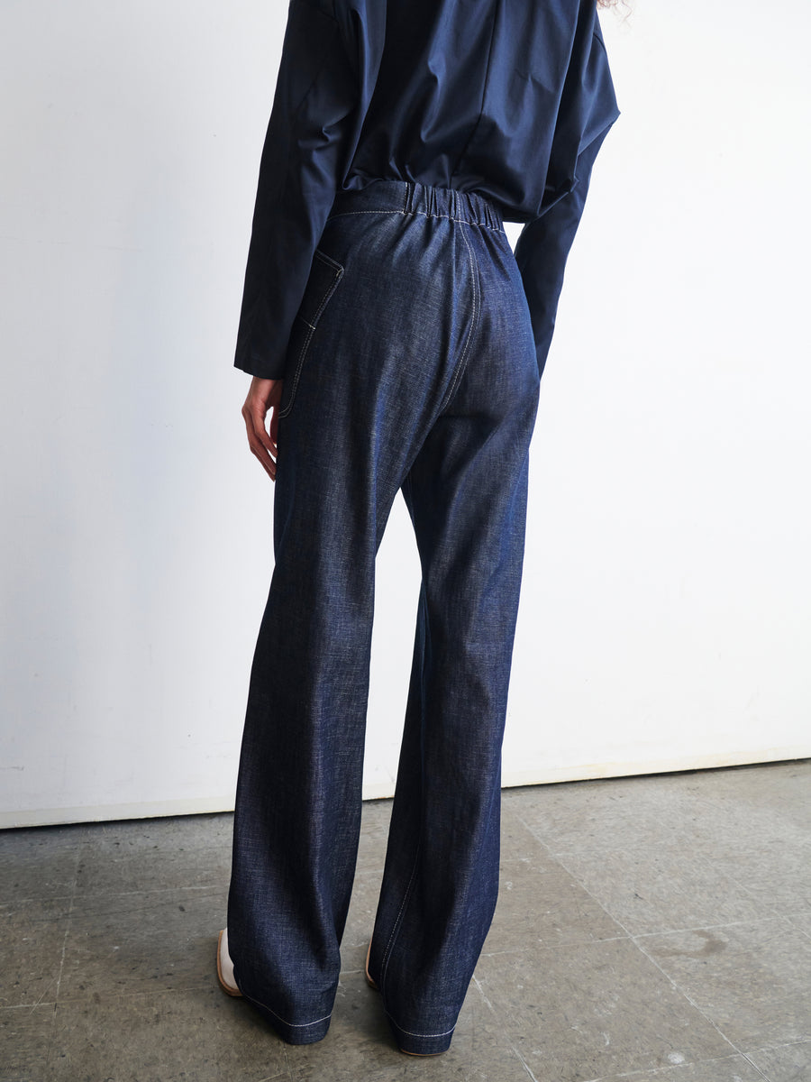 In a minimalistic setting with a concrete floor and white wall, an individual is standing while wearing a dark long-sleeve shirt and the Eda Pant from Zero + Maria Cornejo. These loose-fitting jeans, crafted from GOTS-certified organic cotton, showcase visible seams and pockets. The person's face is not visible.