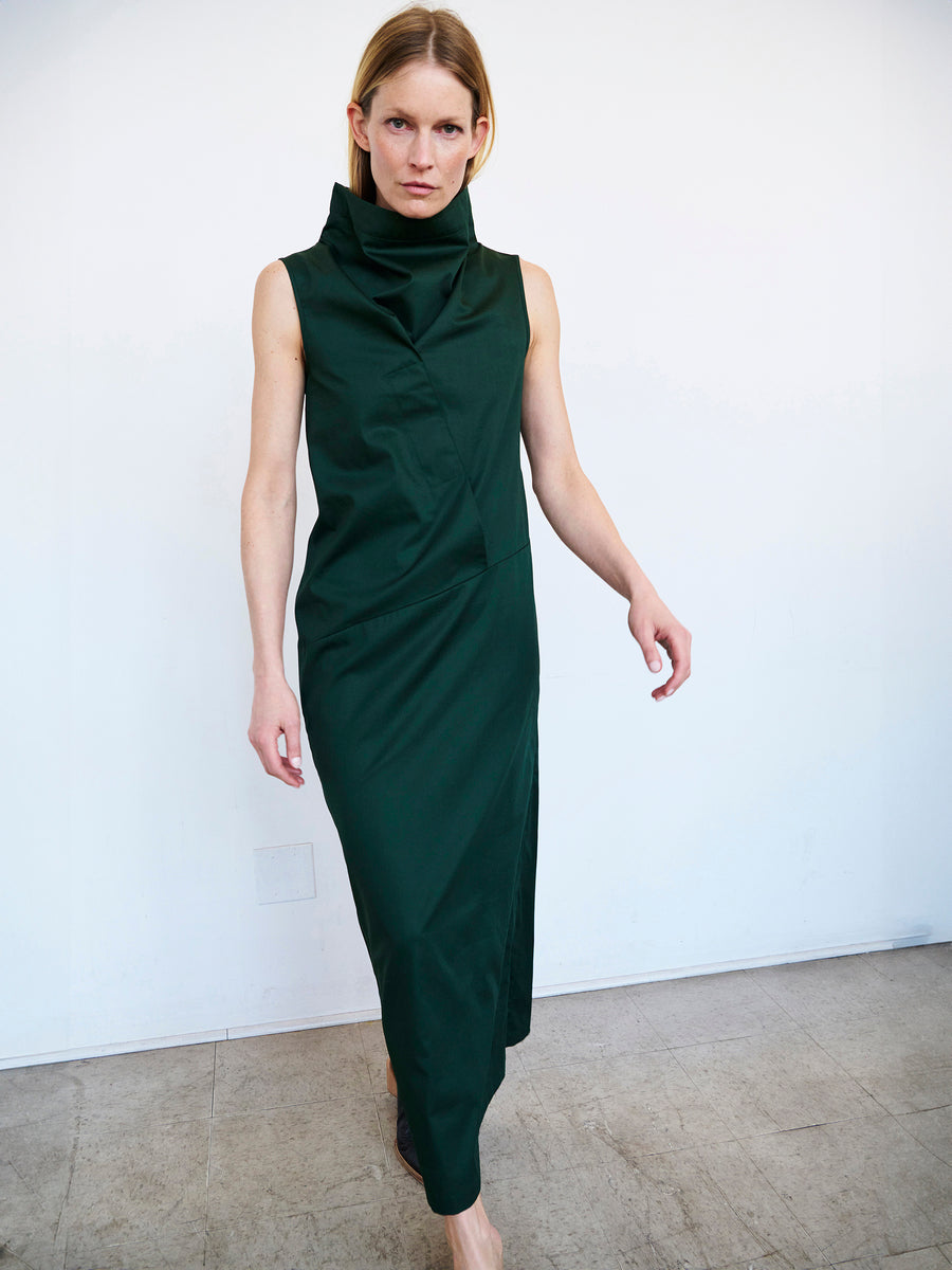 A person with long blonde hair is modeling the Zero + Maria Cornejo Long Fold Dress in dark green. The sleeveless mid-length dress has a draped high neckline and an overlapping design at the waist. They are walking forward in a minimalist setting with a plain background.