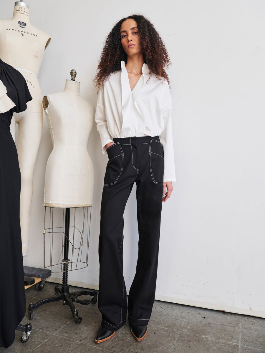 In a studio setting, a person with curly hair exudes confidence while wearing a white shirt and the Eda Pant from Zero + Maria Cornejo, made from organic denim with prominent stitching. Behind them, mannequins display garments made of GOTS-certified cotton, evoking a sense of fashion or design.