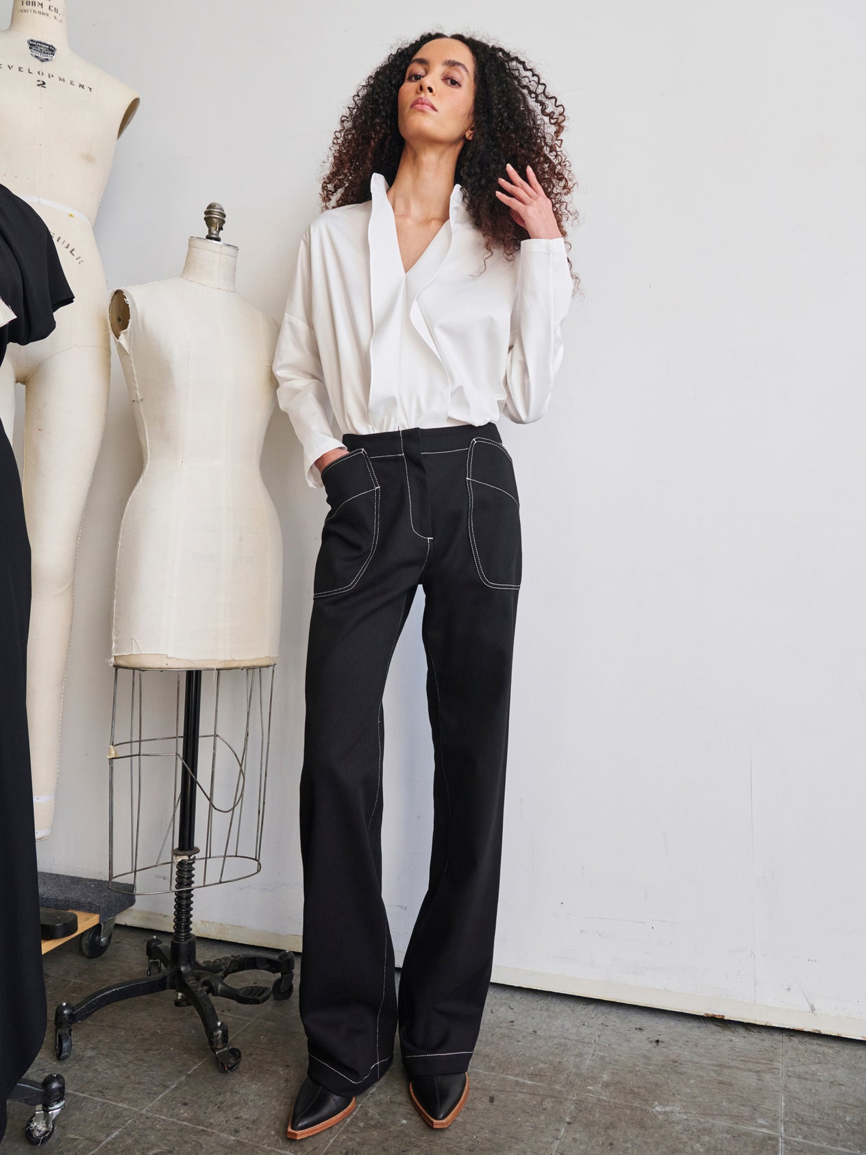 A person with long curly hair is posing in a stylish dark blue top and Eda Pant from Zero + Maria Cornejo. They are standing next to a dress form in a minimalist room with a neutral-colored wall and floor.
