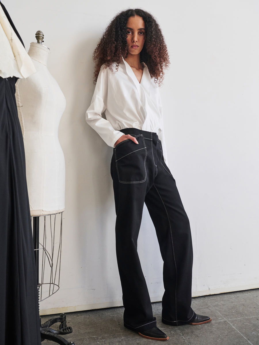 A person with curly hair models a white long-sleeve shirt and the Eda Pant by Zero + Maria Cornejo, leaning against a wall. A dress form and draped fabric in the background create a fashion studio setting that highlights the Cradle to Cradle Certified™ Gold standards.