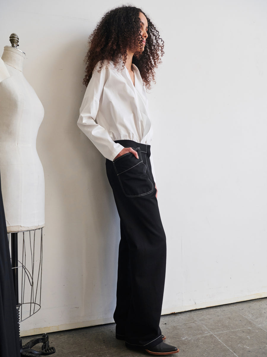 A person with curly hair stands sideways, hands in the pockets of their Eda Pant by Zero + Maria Cornejo, crafted from organic denim and paired with a white shirt. They are positioned next to a dress form mannequin against a plain white wall.