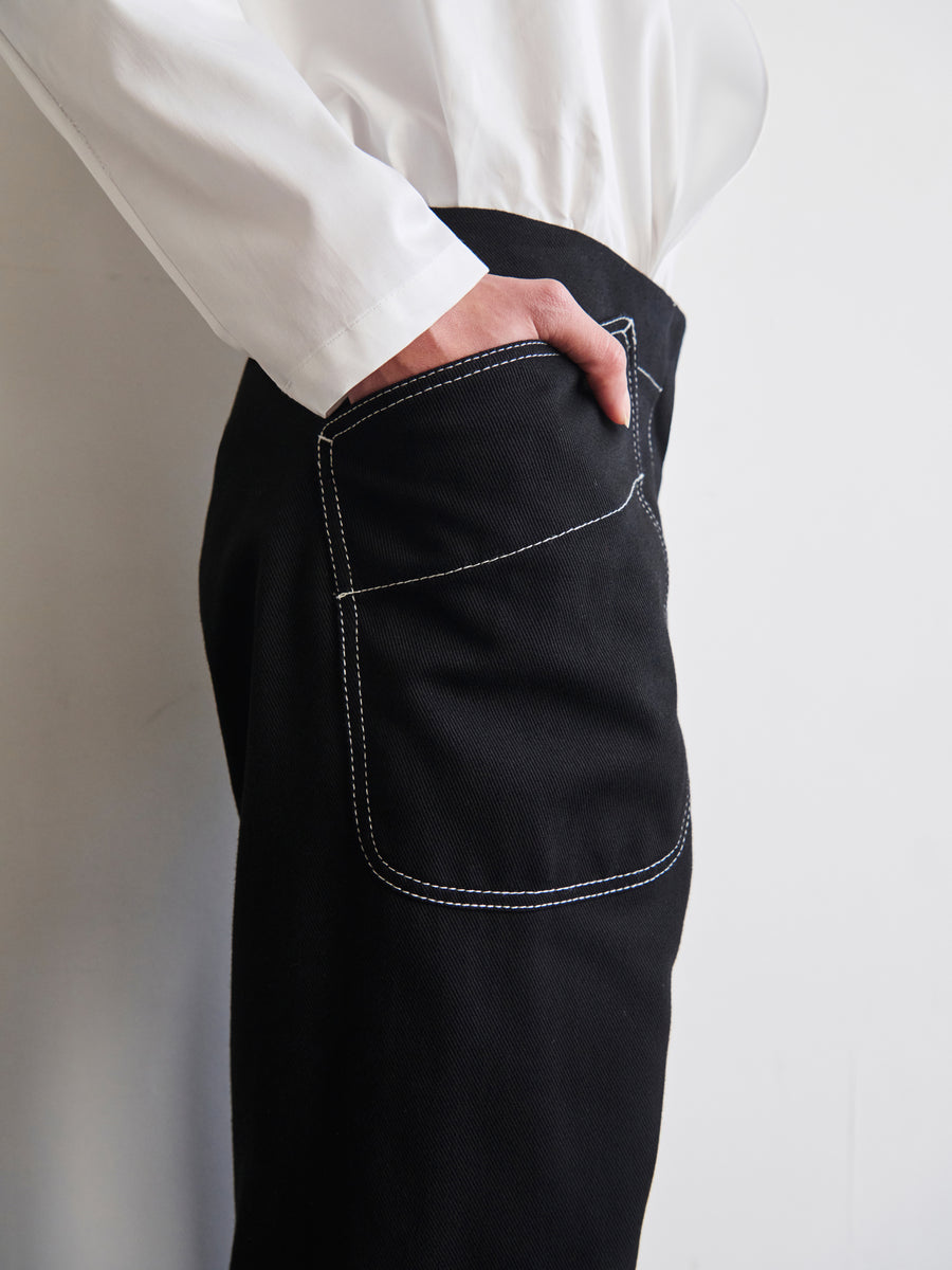 A person wearing a white shirt and the Eda Pant by Zero + Maria Cornejo, characterized by its black organic denim and contrast stitching, is standing with their hand in the back pocket against a plain background.