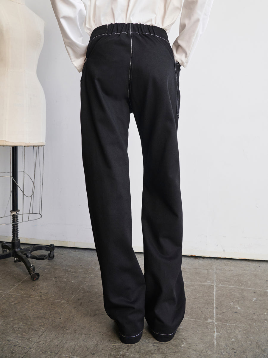 A person wearing the Eda Pant by Zero + Maria Cornejo, crafted from organic denim in a high-waisted black design, stands with their back to the camera. A white mannequin is partially visible to the left, and the floor is gray. The individual complements their outfit with a crisp white shirt.