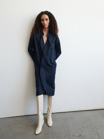 A person with long, curly hair stands against a plain white wall, dressed in a Long-Sleeved Fin Gaban Dress by Zero + Maria Cornejo and wearing white knee-high boots. Their hands rest in the dress pockets, and they maintain a neutral expression.