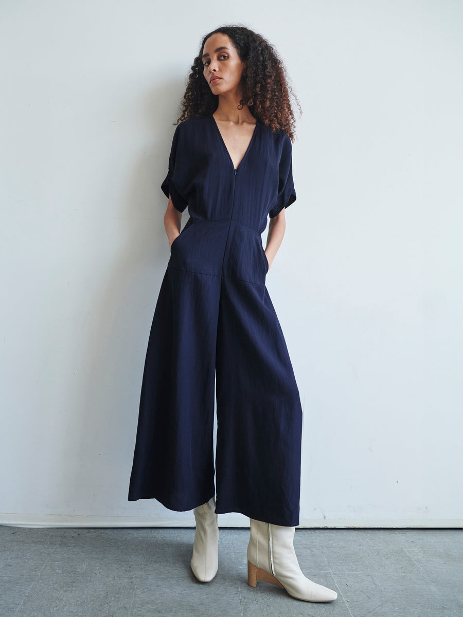 A person with long curly hair is standing against a plain white wall, dressed in the Aissa Eve Jumpsuit by Zero + Maria Cornejo paired with white boots. They have their hands in their pockets and are gazing to the side.