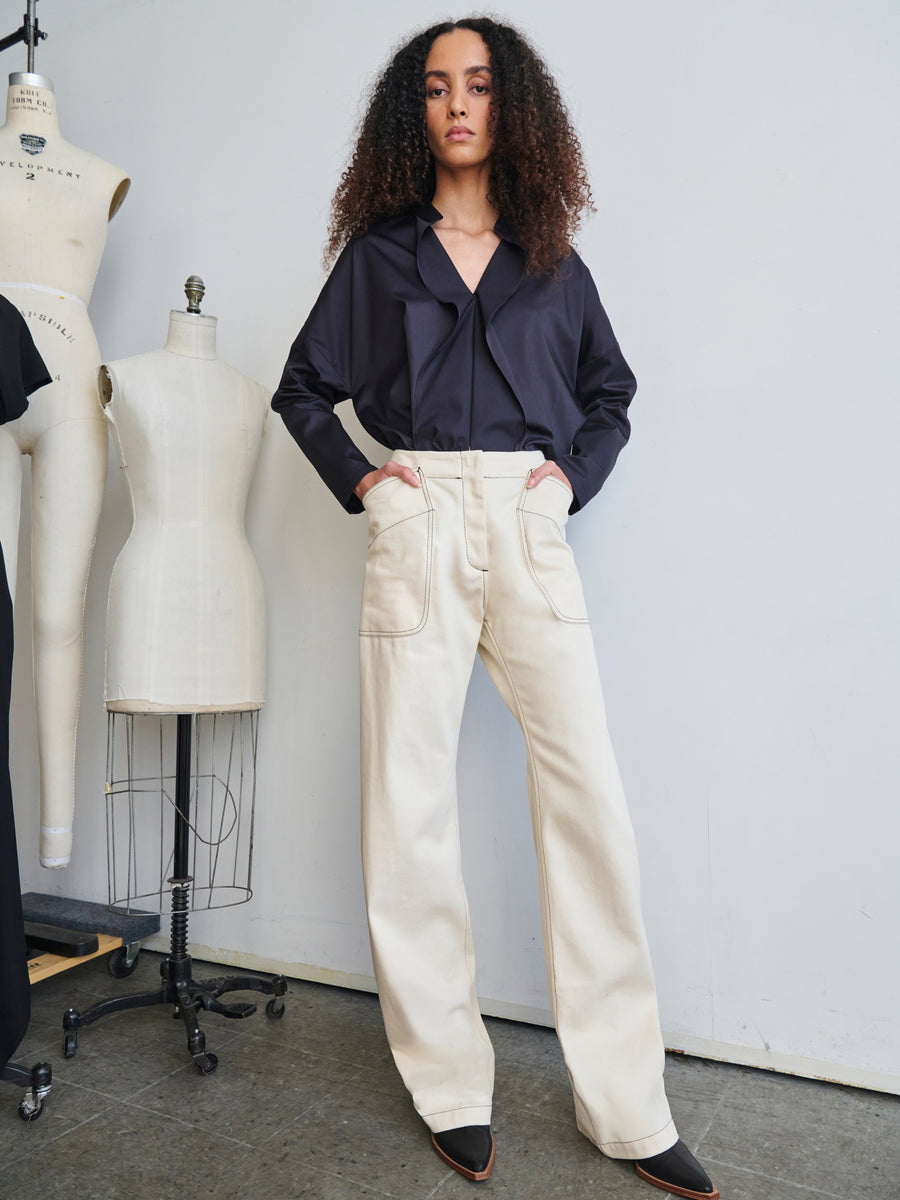 A person with curly hair stands confidently in a fashion studio, wearing a dark long-sleeve blouse and the high-waisted Eda Pant from Zero + Maria Cornejo. Tailors' mannequins are visible in the background, celebrating sustainable style with Cradle 2 Cradle Certified materials.