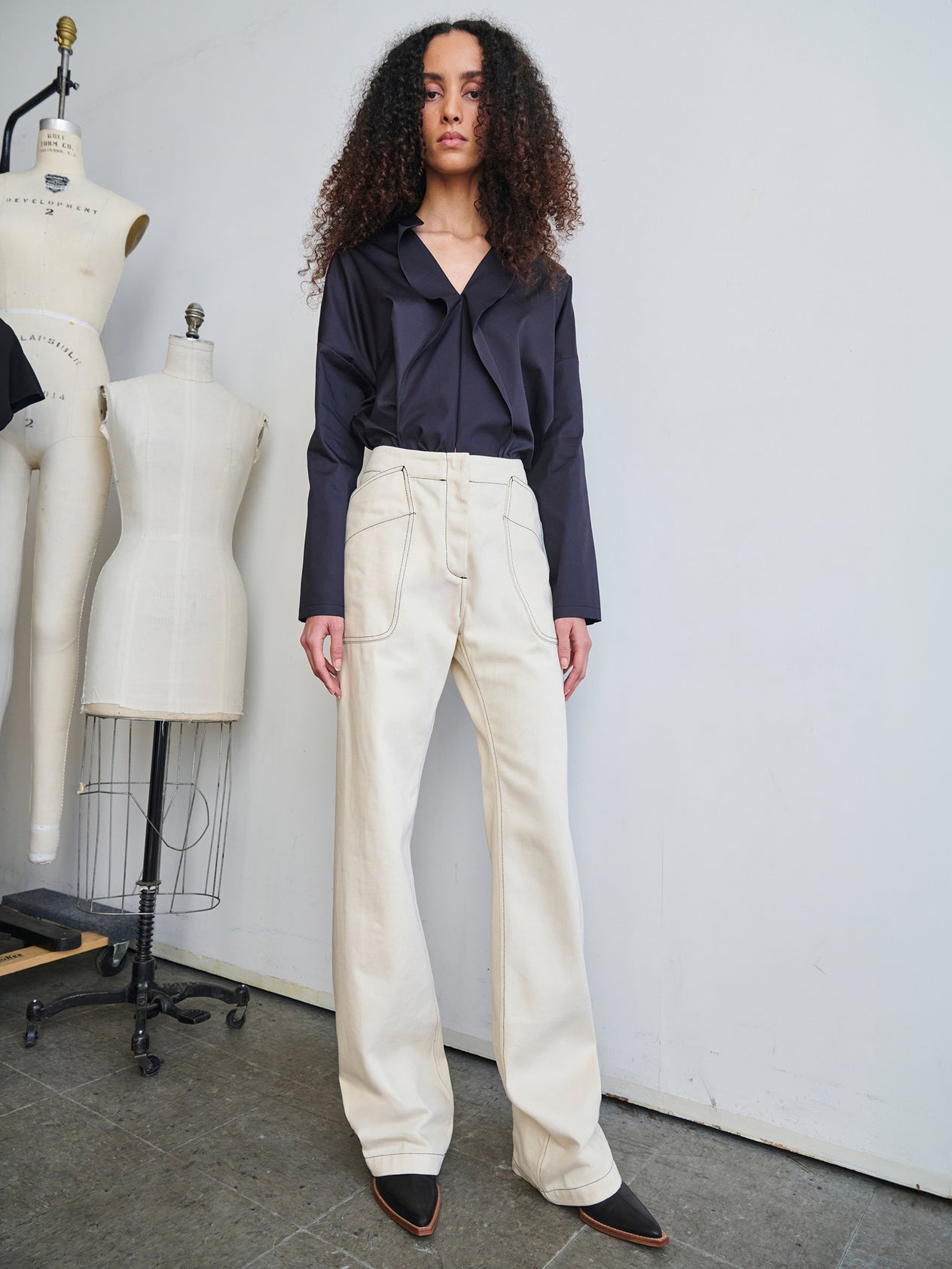A person with long curly hair is posing in a stylish dark blue top and Eda Pant from Zero + Maria Cornejo. They are standing next to a dress form in a minimalist room with a neutral-colored wall and floor.