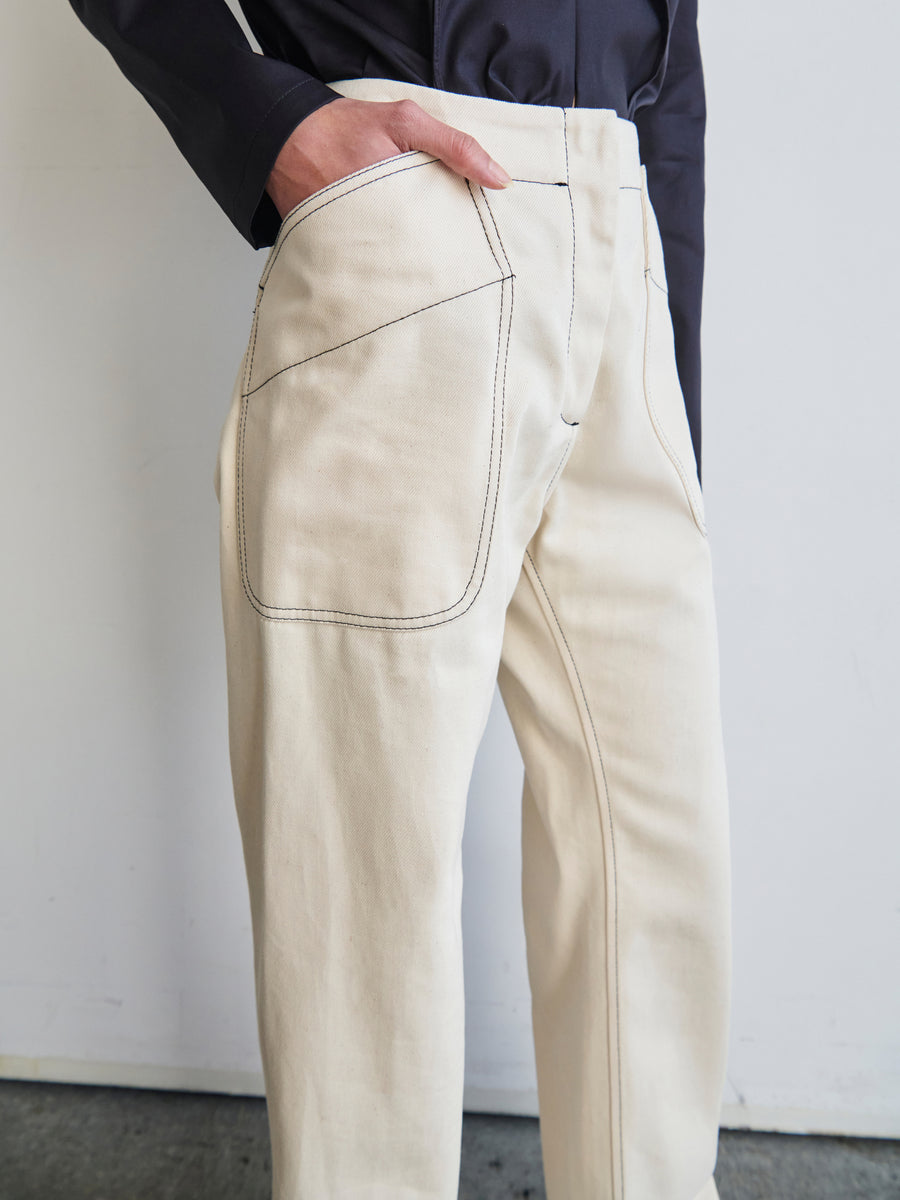 A person is wearing the Eda Pant from Zero + Maria Cornejo, featuring beige fabric with large, visible pockets and black stitching, crafted from GOTS-certified cotton. Their hand rests in the right pocket against a plain white wall, paired with a black long-sleeve shirt.