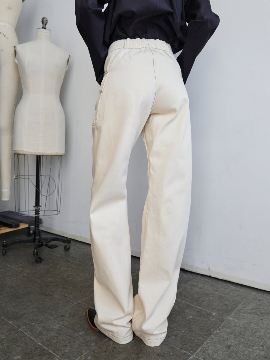 Someone is standing in a room with two dress forms, wearing the Eda Pant by Zero + Maria Cornejo, made from light-colored organic denim. They are facing away, and the floor consists of dark tiles.