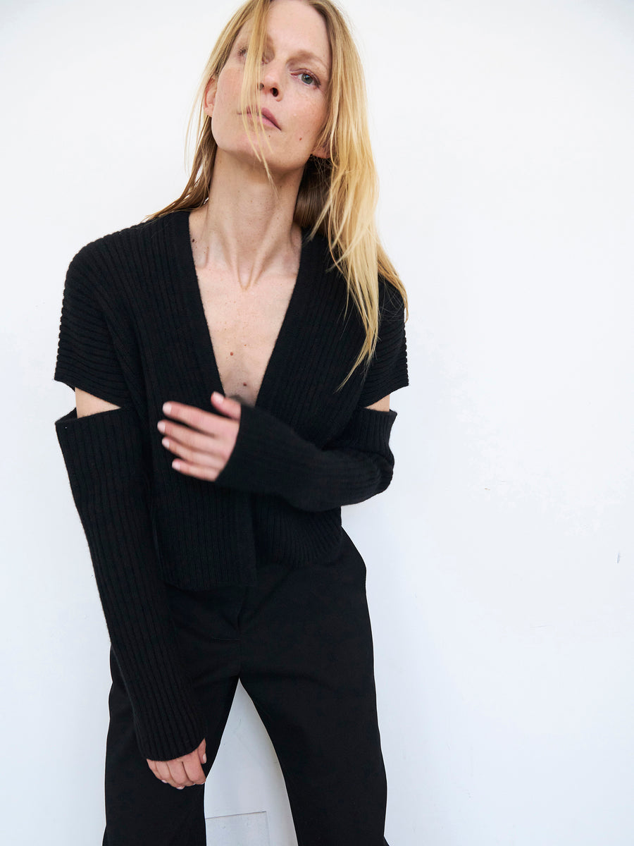 A person with long blonde hair stands against a white background, wearing the Slash Ani Cardigan by Zero + Maria Cornejo. The black ribbed knit design features cut-out shoulders paired with black pants. They have a slight lean and gaze directly at the camera with a neutral expression. The recycled cashmere of the cardigan adds an element of sophistication to the look.