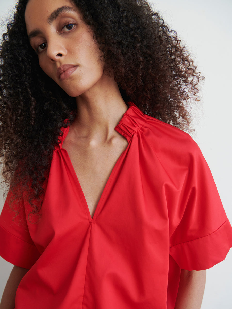 A person with curly hair wearing a red Malia Shirt by Zero + Maria Cornejo, featuring an oversized fit and V-neckline, looks directly at the camera. This year-round shirting style is accentuated by gathered detailing at the shoulders. The backdrop is plain and light-colored.