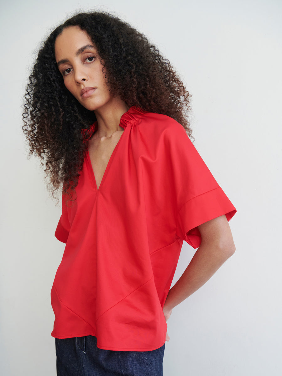 A person with curly hair wears an oversized Malia Shirt in vibrant red, featuring a deep V-neck and gathered collar, paired with dark pants. They stand against a plain white background, looking directly at the camera, embodying Zero + Maria Cornejo's timeless year-round shirting style.