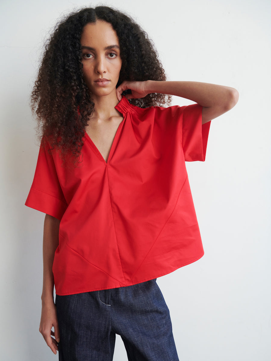 A person with long, curly hair is seen wearing the oversized Malia Shirt in red cotton broadcloth by Zero + Maria Cornejo, paired with dark blue jeans. They stand against a plain white background, exuding a relaxed yet confident aura that perfectly encapsulates year-round shirting style.