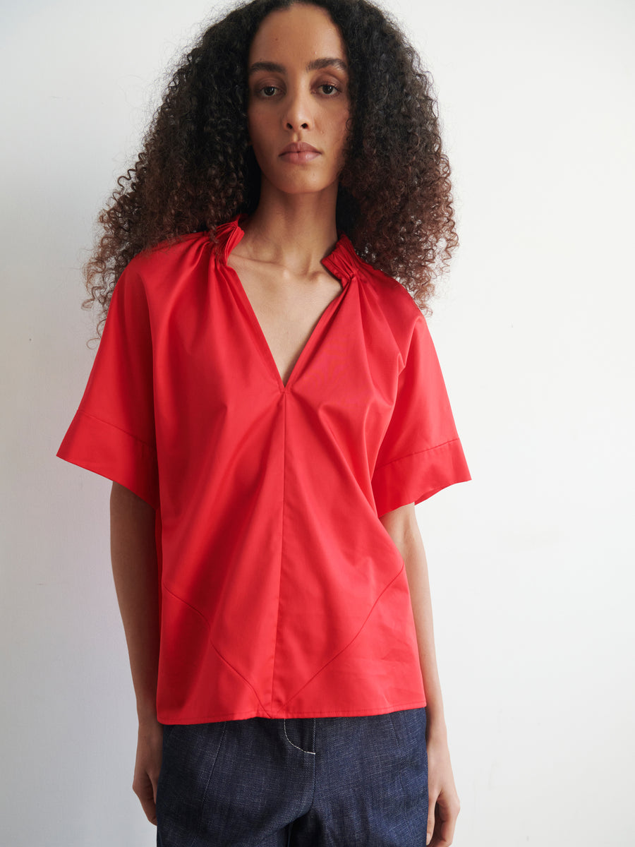 A person with long, curly hair is wearing the oversized Malia Shirt in bright red from Zero + Maria Cornejo. Made from cotton broadcloth, this versatile shirting piece contrasts strikingly against the plain white background as they look directly at the camera.