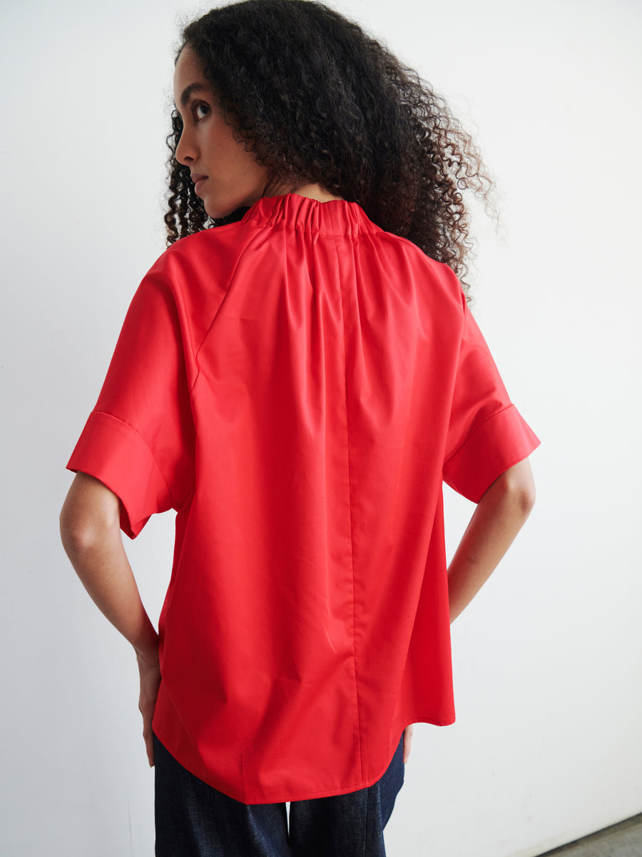 A person with curly hair is wearing the Malia Shirt by Zero + Maria Cornejo, which features a bright red cotton broadcloth fabric, gathered high neckline, and short sleeves in an oversized fit. They are paired with dark pants and face away from the camera against a plain white background.