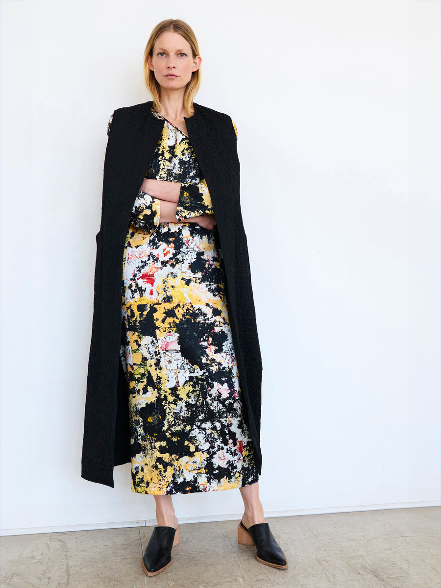 A person with long hair stands against a plain white wall, wearing a colorful abstract-patterned dress and the Luna Waistcoat, a long black sleeveless coat with grosgrain trim from Zero + Maria Cornejo. They also wear black pointed shoes and have their arms crossed while gazing forward.