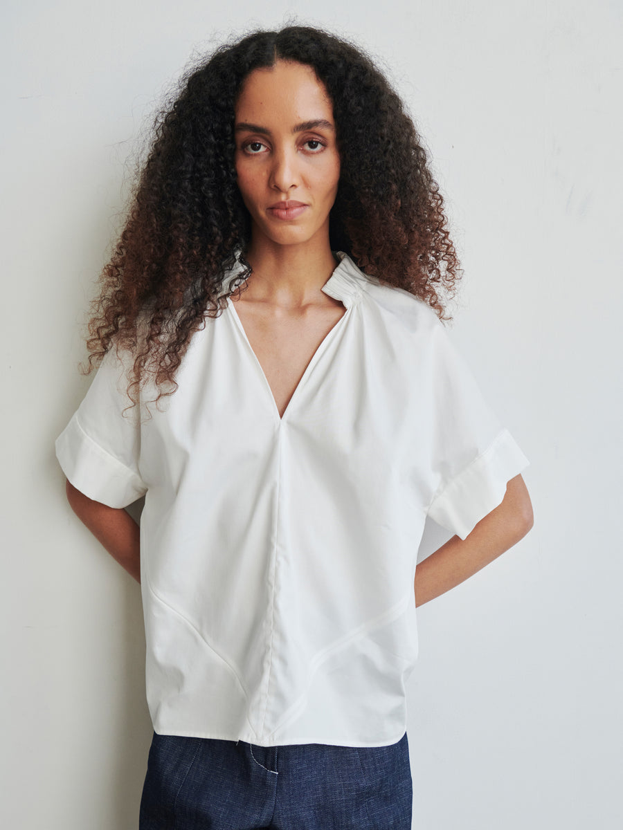 A person with long, curly hair wears the Malia Shirt from Zero + Maria Cornejo, a white, short-sleeved blouse with a v-neckline and an oversized fit. They pair it with dark pants and stand against a plain light-colored background, gracefully positioning their hands behind their back.