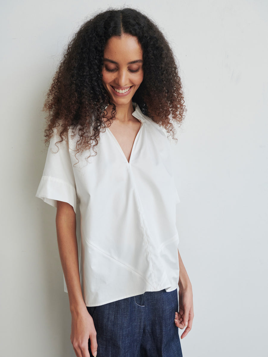 A smiling woman with curly hair stands against a plain wall, wearing the Malia Shirt by Zero + Maria Cornejo paired with dark pants. She looks down with a cheerful expression, embodying the effortless style of year-round shirting.