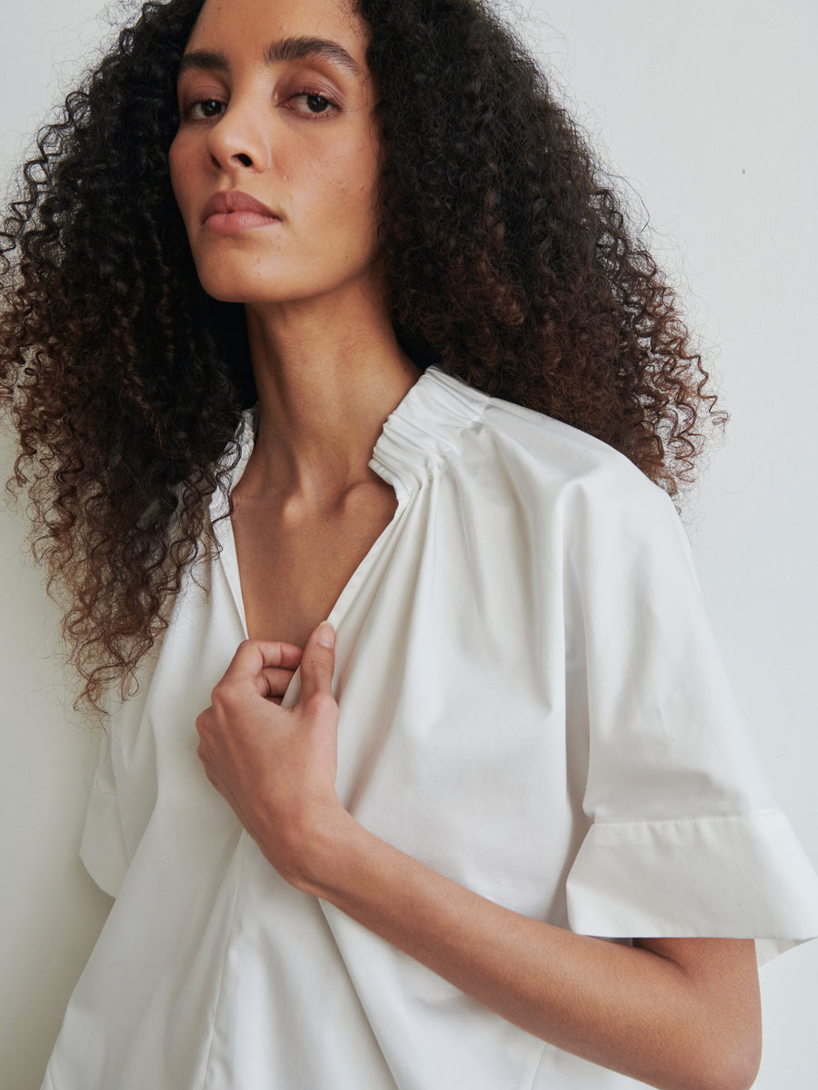 A person with curly hair is wearing the Malia Shirt from Zero + Maria Cornejo, featuring a light color and short sleeves with gathered details at the neckline. The oversized fit of the top adds comfort as they gaze directly at the camera against a plain background.