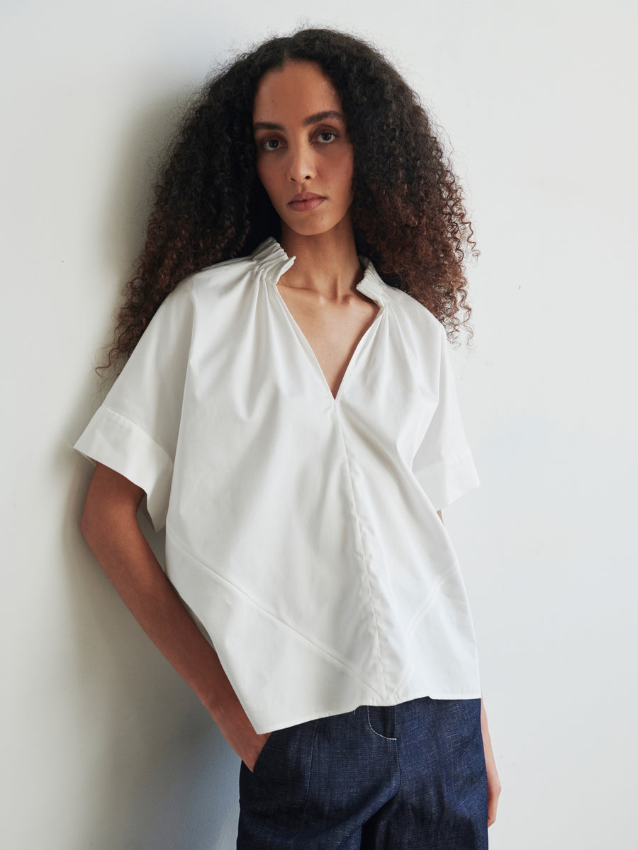 A person with long, curly hair stands against a plain white wall, wearing an oversized Malia Shirt by Zero + Maria Cornejo and dark jeans, gazing directly at the camera.