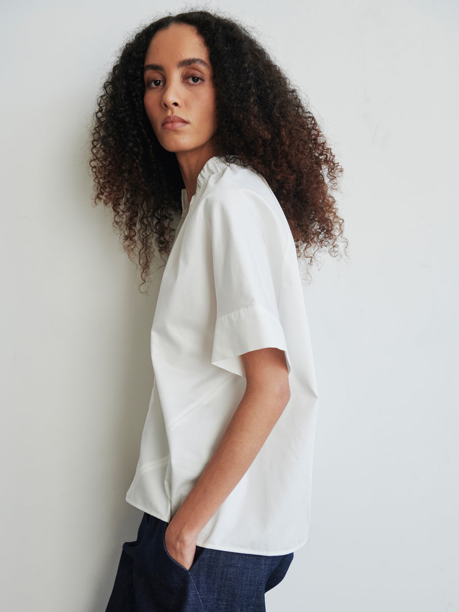 A person with curly hair is wearing an oversized Malia Shirt by Zero + Maria Cornejo, paired with dark pants while standing sideways against a plain white background. Their hands are in their pockets, exuding a neutral expression.