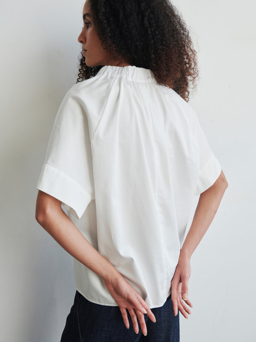 A person with curly hair is seen from the back, wearing the Malia Shirt by Zero + Maria Cornejo in an oversized fit, paired with dark pants. They stand against a plain light-colored wall, their hands resting on their lower back.