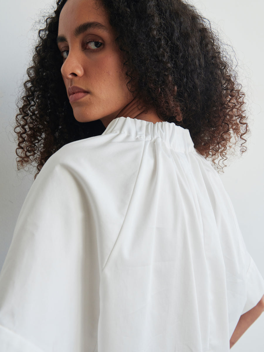 A person with curly hair is wearing the oversized Malia Shirt by Zero + Maria Cornejo, made of cotton broadcloth and featuring a gathered neckline. They are looking over their shoulder at the camera against a plain background.